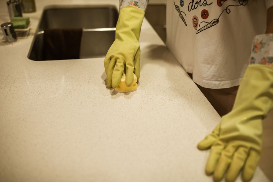 Close-up of a person wearing yellow rubber gloves and a white apron with a floral pattern, actively cleaning a smooth, light-colored kitchen countertop using a yellow sponge. In the background, a stai