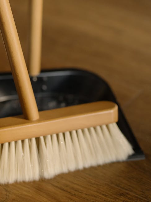 Close-up view of a wooden-handled broom with beige synthetic bristles, set on a polished wooden floor. The background shows a portion of a black dustpan, and the lighting highlights the clean, smooth 